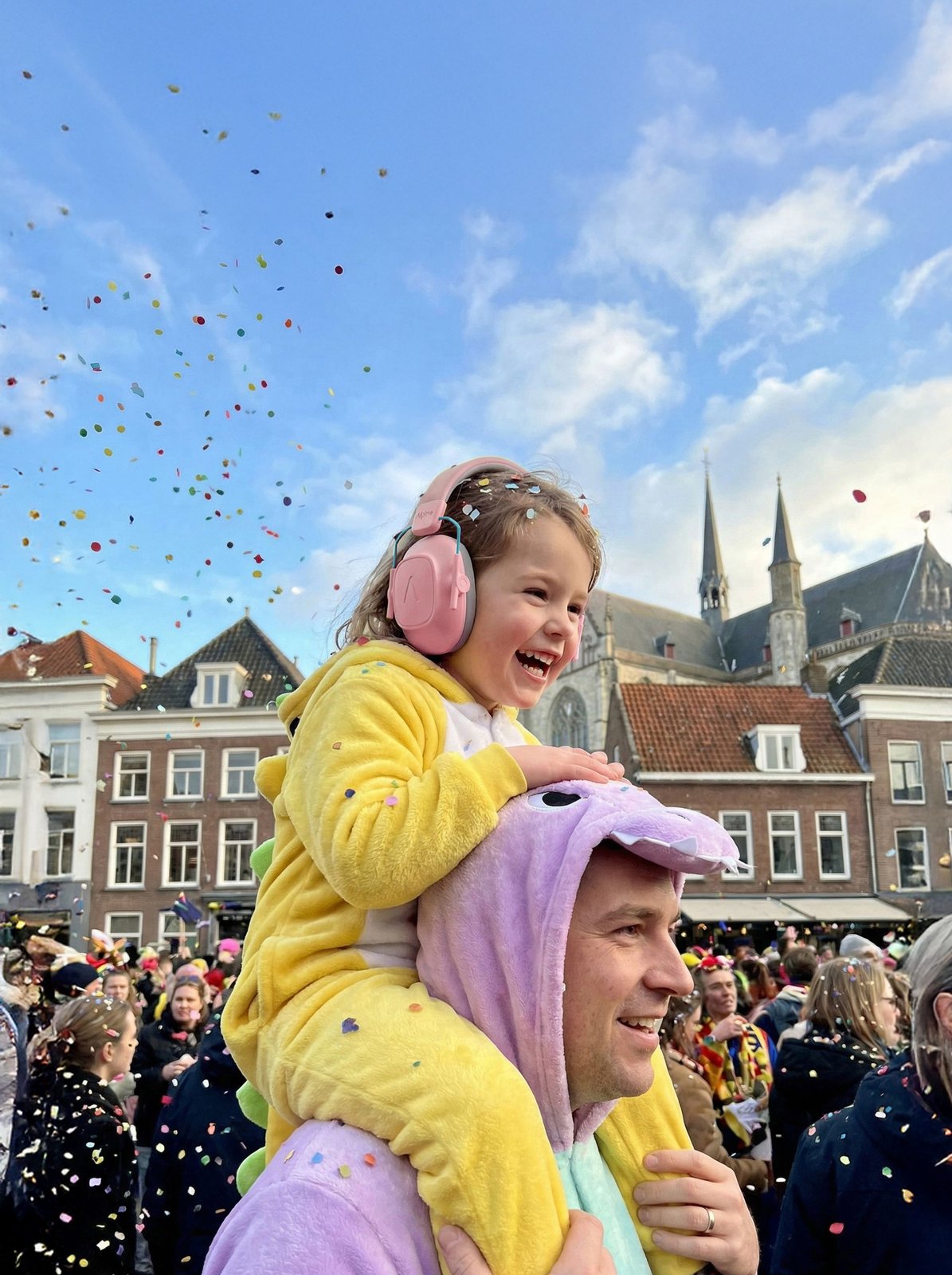 Girl in a yellow dinosaur onesie wearing pink Alpine earmuffs under falling confetti at Carnaval